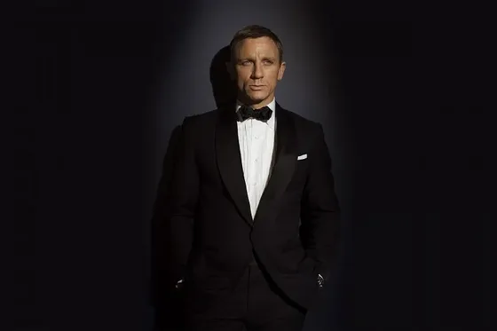 A man in a black tuxedo stands against a dark background.