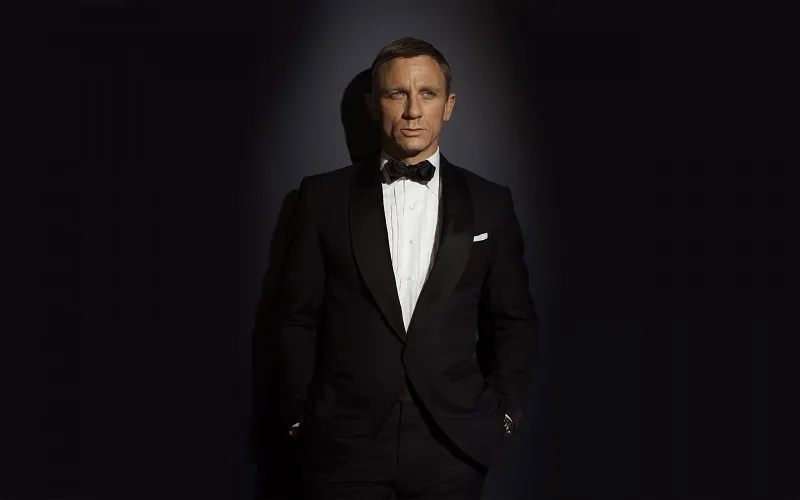A man in a black tuxedo stands against a dark background.